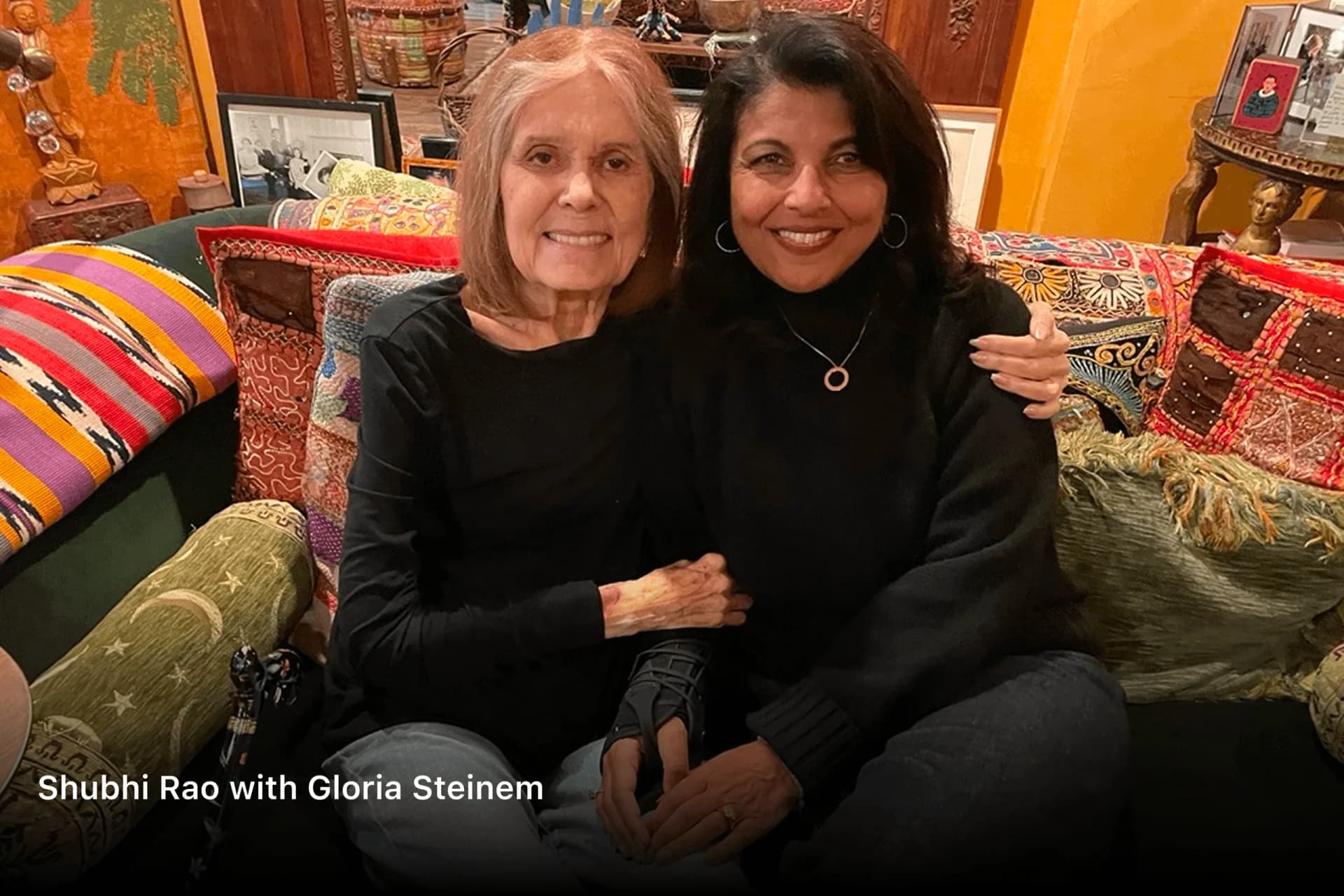 Shubhi Rao seated with Gloria Steinem on a couch of embroidered cushions